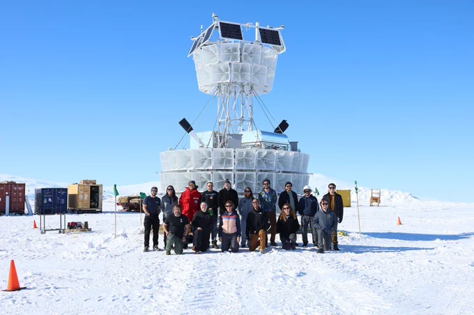 A group of people pose in the snow in front of a large white scientific instrument with solar panels, set against a bright blue sky and snowy landscape. Shipping containers and equipment are visible in the background.