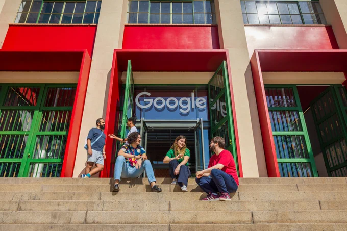 A group of five people sit and chat on the outdoor concrete steps in front of a modern building with large red and green framed doorways and a Google logo.