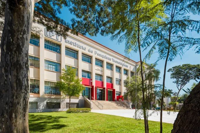 A wide shot of the exterior of a large, multi-story white building labeled "Instituto de Pesquisas Tecnológicas" with a green lawn and trees in the foreground.
