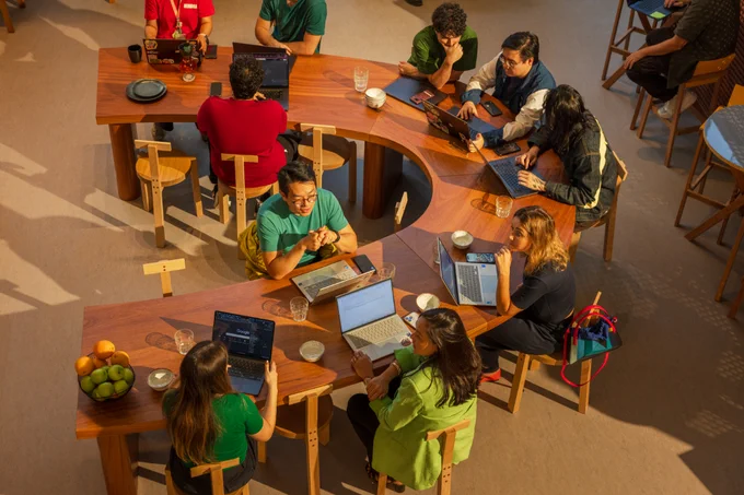 An overhead view of a group of people sitting along a winding, S-shaped wooden table working on their laptops.
