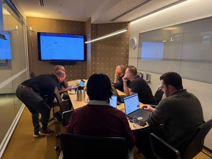 A trainer assists a participant at their laptop during an active workshop, with a large screen in the background displaying a software dashboard.