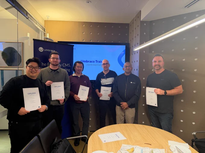 A group of six men posing in a meeting room while holding their diplomas after successfully completing the "Umbraco Training Cloud Developer Course."