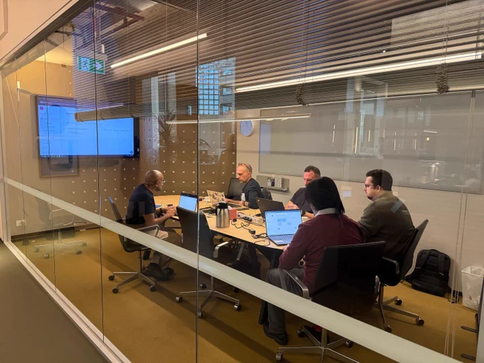 A group of professionals viewed through a glass office wall, sitting around a long table with laptops during a technical training session.