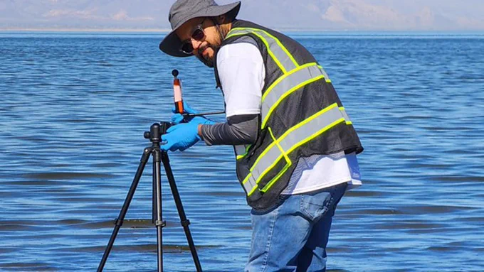 A person wearing a wide-brimmed hat, reflective vest, and gloves is standing in shallow water next to a tripod with a mounted instrument. The background features a large body of water and distant mountains under a clear sky.