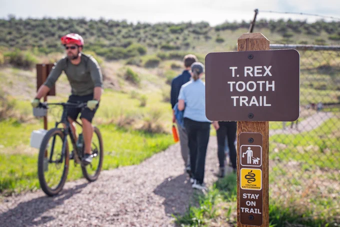 Focused shot on a sign that says "T. rex Tooth Trail". In the background is a person riding a bike and some people hiking.