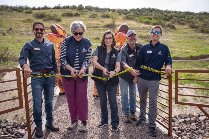 A group of people cutting a ribbon with two costumed dinosaurs in the background. 