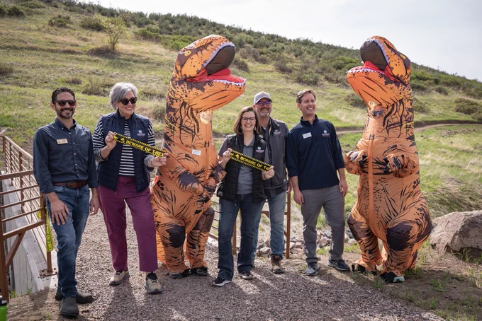 Group of people with cut ribbon, standing with costumed dinosaurs. 
