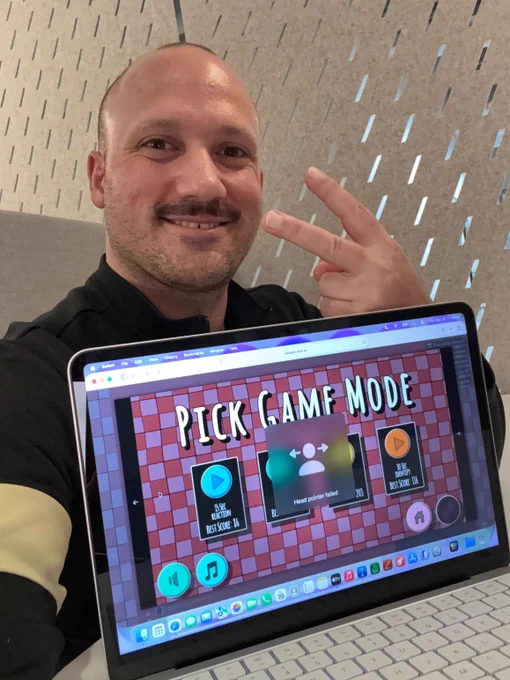 A smiling man takes a selfie while holding up an open laptop displaying a screen for an accessibility challenge game, while giving a peace sign.