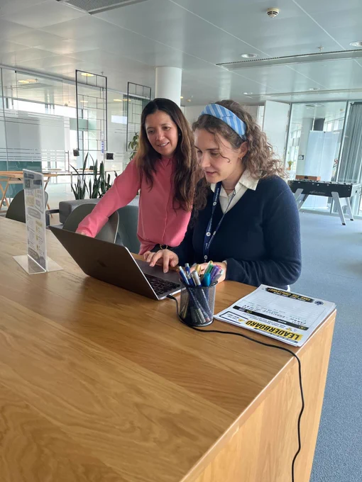 Two women work together at a laptop on a standing desk in a bright, open office. One wears a Siteimprove lanyard. A printed leaderboard sheet for the accessibility challenge is visible next to the computer.