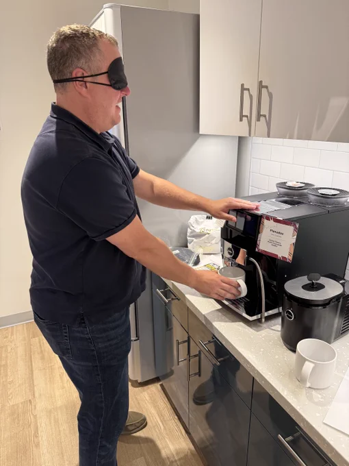 A man wearing a black sleep mask stands at a coffee machine in a modern office kitchen, reaching for the machine by touch while holding a white mug in his other hand.