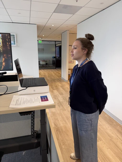 A woman with her hair in a bun and a Siteimprove lanyard stands at a standing desk with a laptop and printed challenge instructions, appearing to interact with the screen without using her hands.