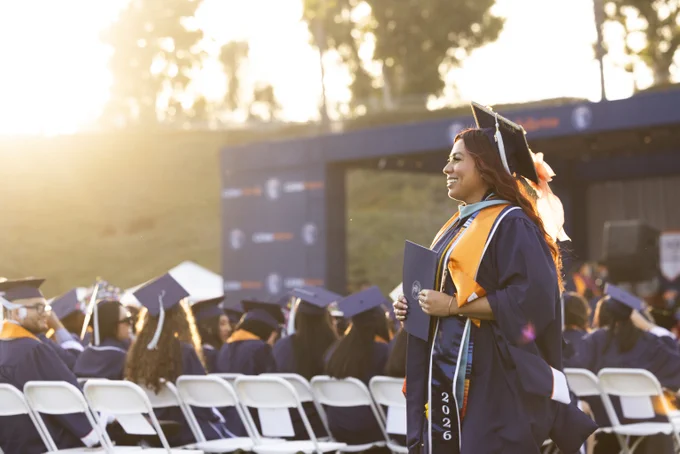 A graduate in a cap and gown stands smiling, holding a diploma cover, at a university commencement ceremony. The background shows rows of seated graduates in similar attire, with trees and a stage in the distance. The sunlight creates a warm atmosphere.