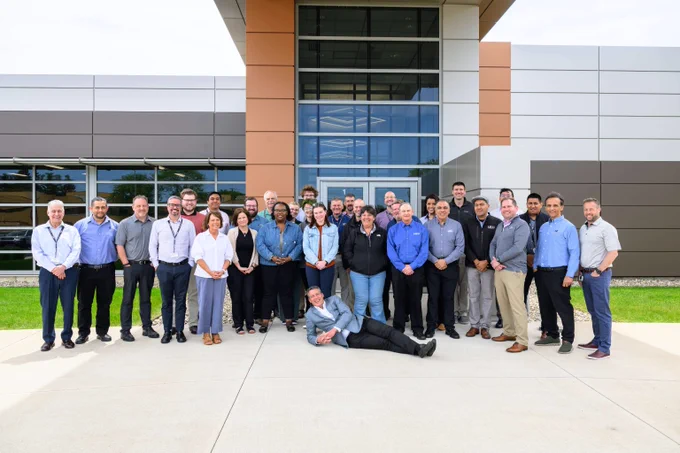 Group of 27 diverse professionals posing outside a modern office building on a sunny day, with one person lying playfully in front.