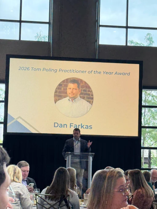 Dan Farkas speaks while standing at a podium in front of a projector screen that reads "2026 Tom Poling Practitioner of the Year Award" at the top. His photo is in a circle frame in the middle and his name is below it. Some people seated at the event can be seen sitting before the stage where Dan Farkas is speaking. 