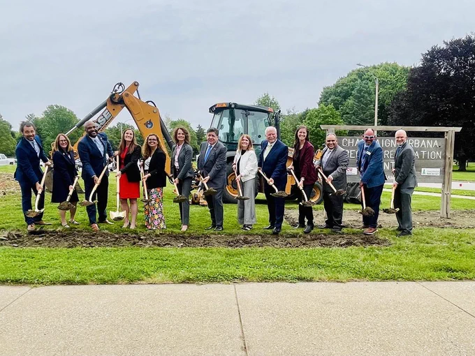 A group of people in business attire holds shovels at a construction site. A yellow excavator is in the background.