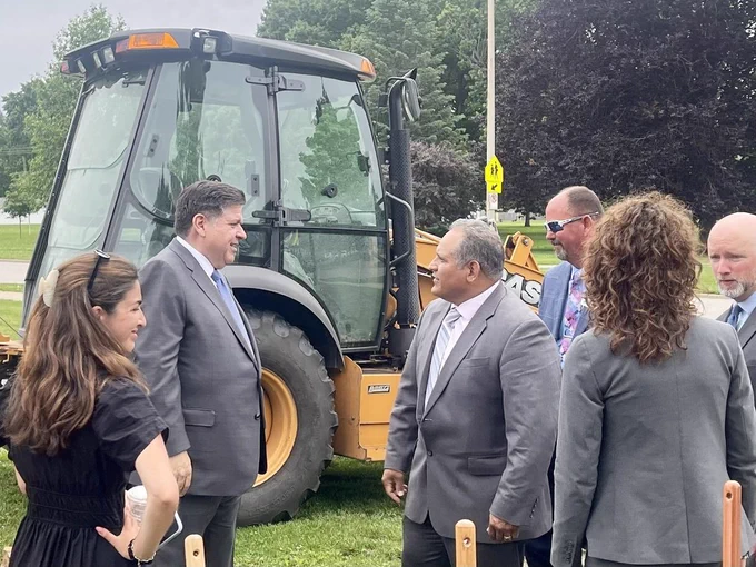 Five people in suits stand near a yellow bulldozer on a grassy area with trees.