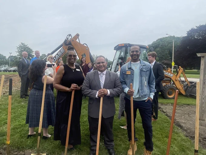 Four people stand on a grassy area holding shovels. Two men and two women, with a machinery vehicle in the background.