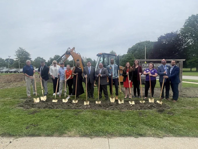 People hold shovels at a construction site, with a machinery vehicle in the background.