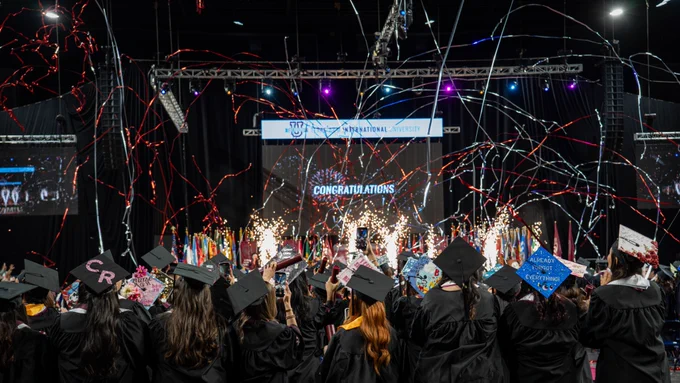 A crowd of graduates facing the stage with a screen that says "Congratulations" with streamers and fireworks.