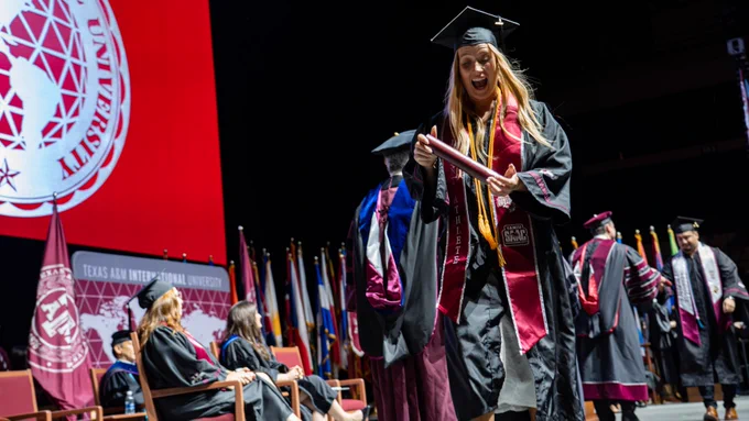 A smiling Texas A&M International Graduate crossing the stage, and looking at their diploma.