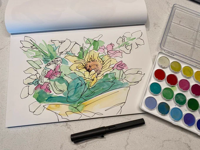 A spread of sketchbook paper, watercolors and a Lamy pen. A completed watercolor painting features a bouquet of flowers in a loose but colorful style. The flowers are abstract with black ink contrasting soft, pastel colors.