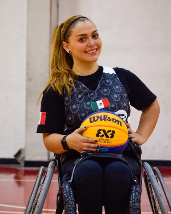A woman in a wheelchair holds a basketball, smiling. She wears a jersey with a Mexican flag.