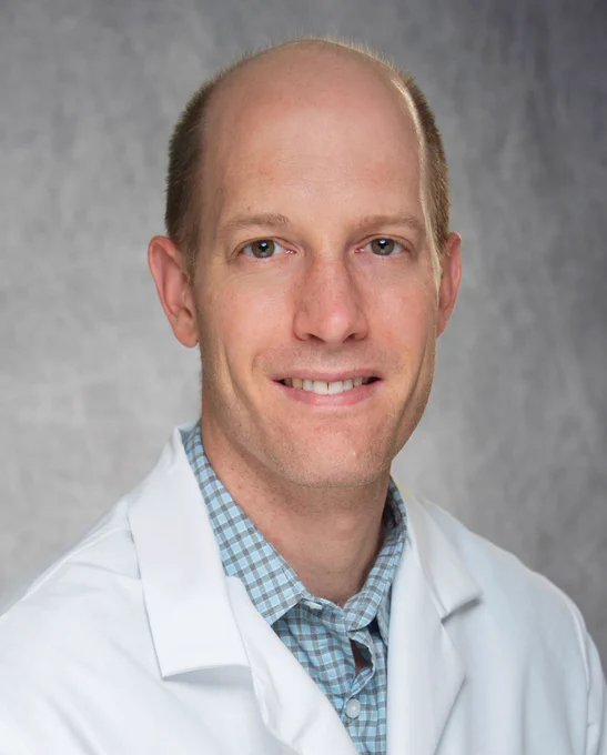 A smiling man with light skin and a shaved head wearing a white lab coat over a blue checkered shirt. The background is a soft gray gradient.