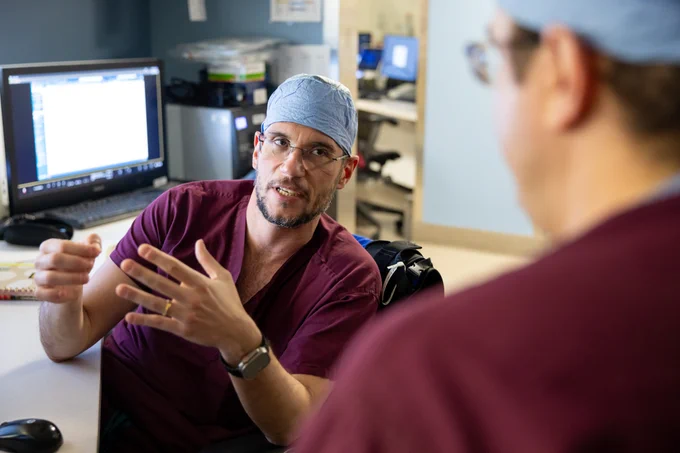 A man wearing a blue surgical cap and maroon scrubs is sitting at a desk, engaged in conversation with another person facing away from the camera. A computer with an illuminated screen is visible in the background. The room appears to be a medical setting.