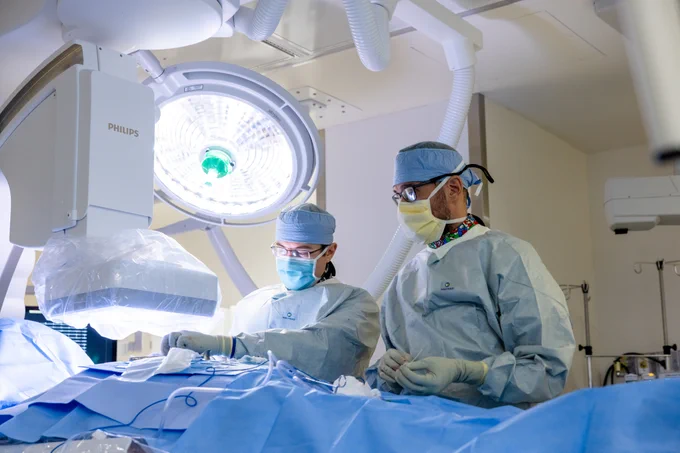 Two surgeons in masks and blue scrubs operate under bright surgical lights in a sterile environment. One adjusts surgical instruments, while the other observes closely, with a large C-arm machine and monitor visible in the background.