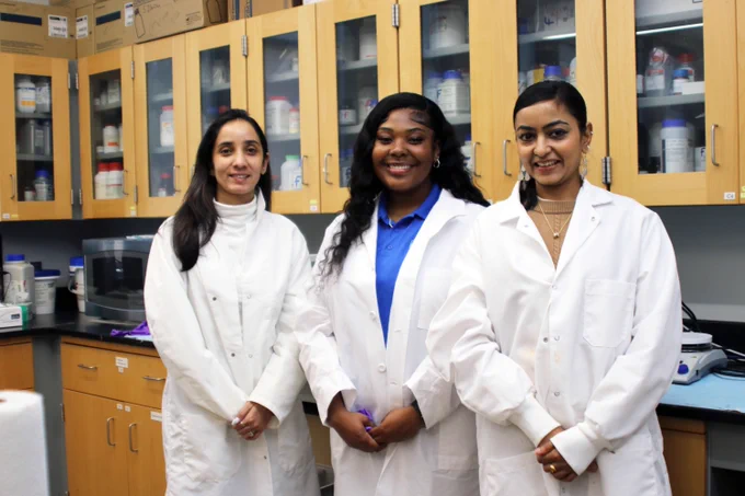 three students wearing lab coats smiling in a campus lab