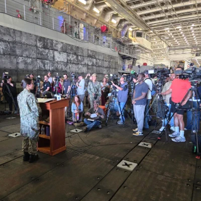 Johnson, with her back to the camera, stands at a lectern speaking to a large crowd of press.