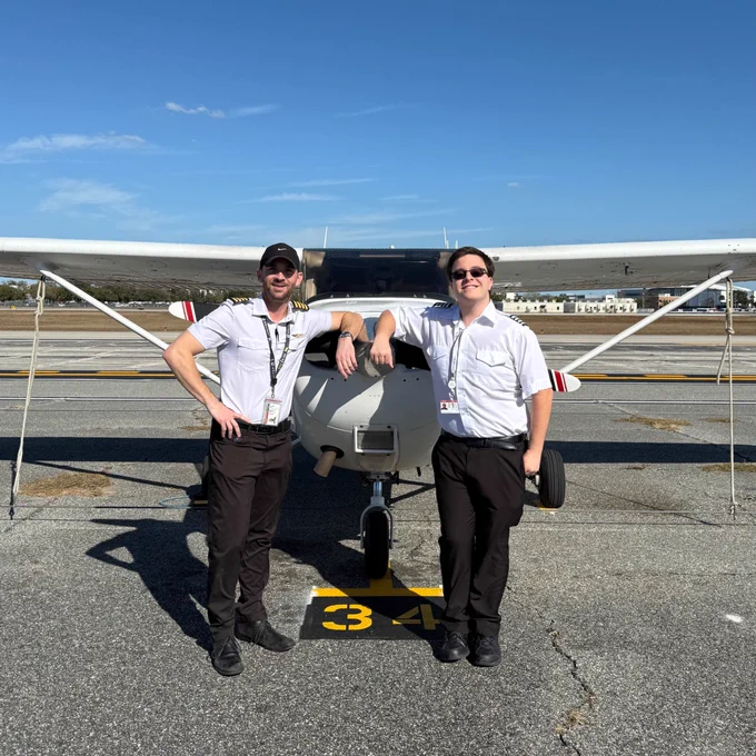 Two individuals standing in front of a small airplane on an airport runway. Both are wearing pilot uniforms with sunglasses. The sky is clear.