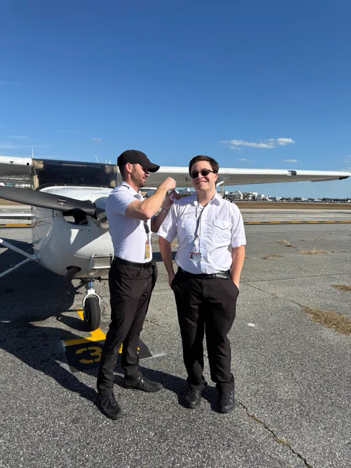 Two people are standing on an airport tarmac beside a small aircraft. They are both wearing aviation uniforms and sunglasses. A runway and clear blue sky are visible in the background.