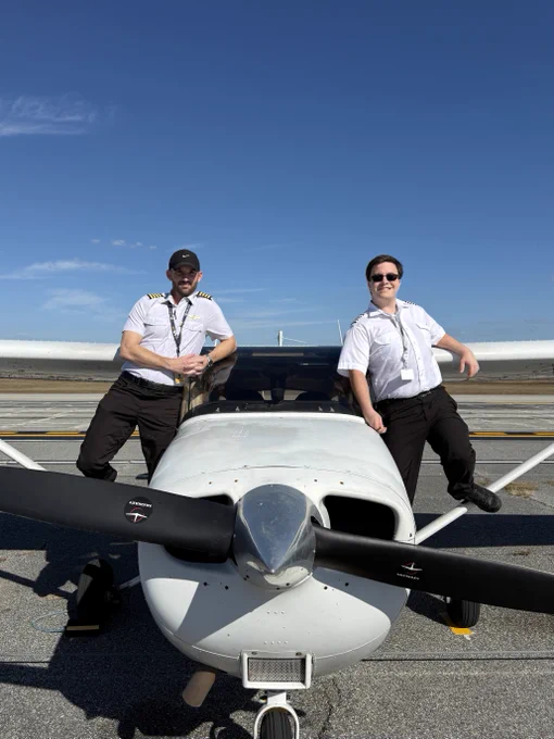 A small aircraft is parked on a runway under a clear blue sky. Two people in pilot uniforms are posing confidently, one leaning against each wing.