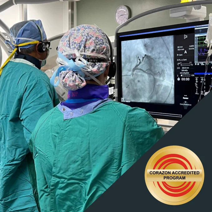 Medical staff are in a hospital room focused on a large medical screen displaying an angiogram. The staff wear protective gowns, head coverings, and headsets. A badge with the text "Corazon Accredited Program" is shown in the lower right corner.