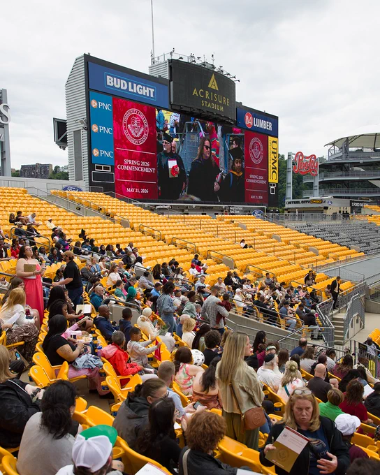 A crowd is seated in the yellow chairs of Acrisure Stadium, viewing a large screen displaying "Spring 2026 Commencement." The screen also shows live footage of individuals participating in a graduation ceremony. Bud Light and 84 Lumber advertisements are visible on either side of the screen.