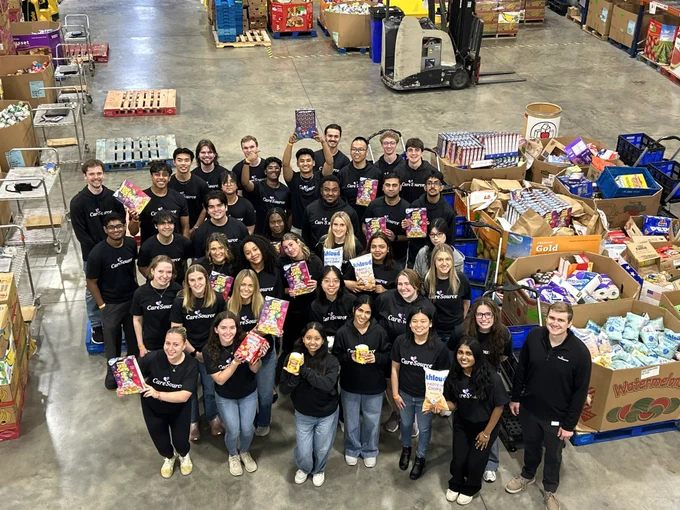 A group of people in matching shirts stands together inside a warehouse filled with assorted food items and supplies. Many are holding various packaged goods. They are smiling and looking up toward the camera. The setting appears to be organized, with products in boxes and on pallets around them.