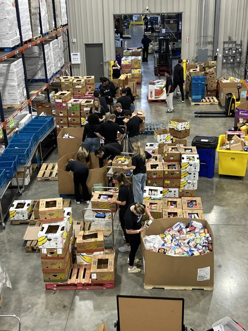 A group of people are organizing and sorting food items in a large warehouse space. There are numerous boxes near several individuals are working. The warehouse has high shelves filled with goods, and pallets scattered around.