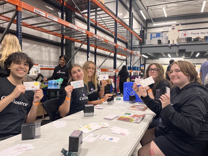 A group of people sitting at a table in a warehouse environment, holding up colorfully decorated cards. They are wearing matching "CareSource" shirts. The setting includes storage racks and a poster with the CareSource logo.