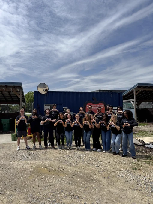 A group of people standing outside, posing in front of a blue shipping container with an apple logo. They are under a partly cloudy sky, and some are making heart shapes with their hands. The scene includes two shelters with metal roofs on either side.