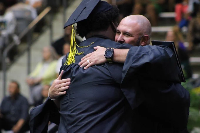 Two people in black caps and gowns hug during an indoor graduation ceremony, with seated guests visible in the background.