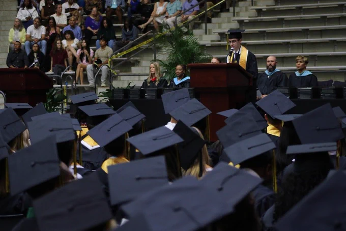 A graduate in a black gown with a gold stole speaks at a podium on a stage while rows of graduates sit facing forward and families watch from the bleachers.