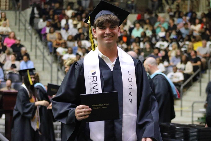 A graduate in a black cap and gown with a white stole reading “LOGAN” and “DRIVEN SCHOOL OF INNOVATION” holds a diploma folder labeled “Robinson High School, Little Rock, Arkansas” in front of a seated audience.