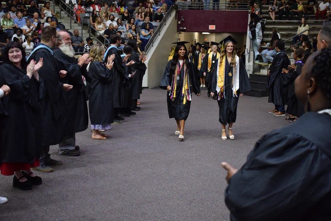 Two graduates in black caps and gowns walk down an aisle lined with clapping faculty members, with more graduates and spectators visible behind them.