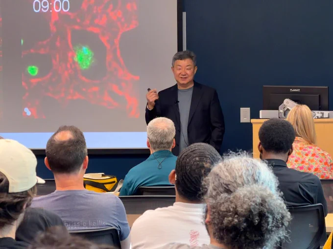 A presenter stands at the front of a seminar room, speaking to a seated audience while holding a small remote. Behind the presenter, a projected slide shows a colorful microscopic image in red and green.