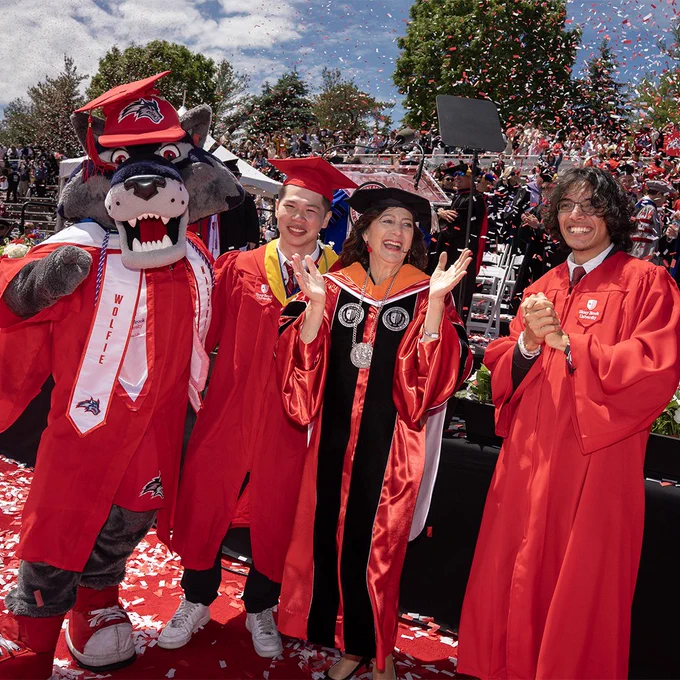 Wolfie stands with graduates and university leadership onstage during Stony Brook University’s commencement ceremony as confetti falls around them.