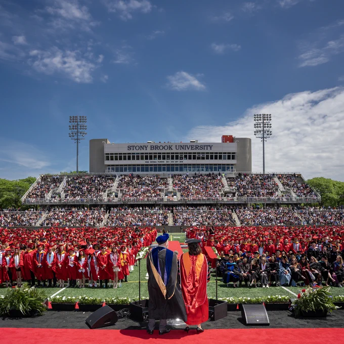 Wide view of Stony Brook University’s commencement ceremony with graduates seated on the field and a full stadium crowd in the background.