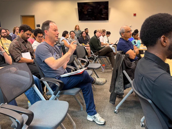 An audience sits in rows of chairs during a lecture, with one attendee holding a microphone and gesturing while asking a question. Other attendees listen, some taking notes or looking toward the speaker.