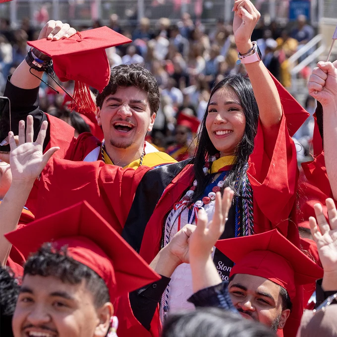 Two graduates in red caps and gowns take a selfie with Wolfie during commencement on the stadium field.