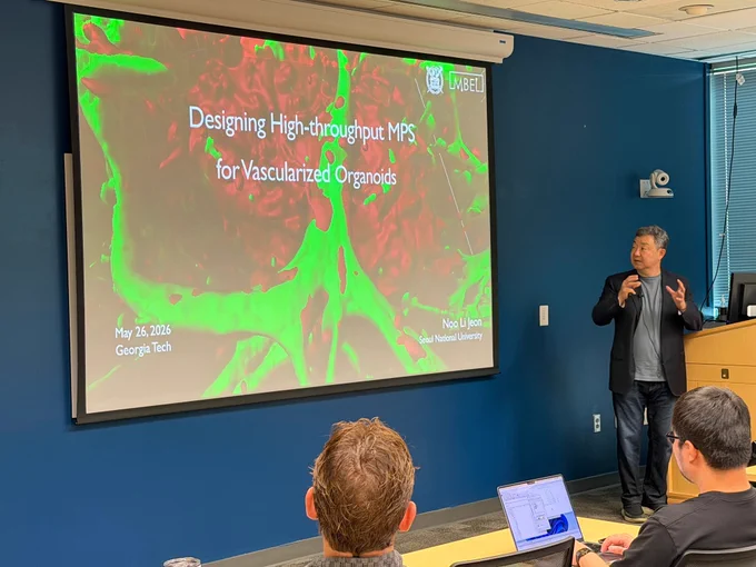 A presenter speaks beside a large projection screen displaying a slide titled “Designing High-throughput MPS for Vascularized Organoids,” with “May 26, 2026, Georgia Tech” visible at the bottom. Attendees sit facing the screen in a seminar room.